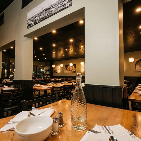 Warmly lit restaurant interior with wooden tables, set with white dishes, and dark chairs.