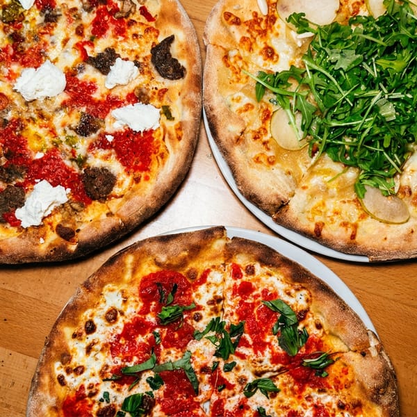Three gourmet pizzas on a wooden table, including a meat and ricotta, a pear and arugula, and a Margherita.