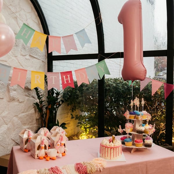A pink-themed first birthday party setup with cake, cupcakes, balloons, and a "HAPPY BIRTHDAY" banner.