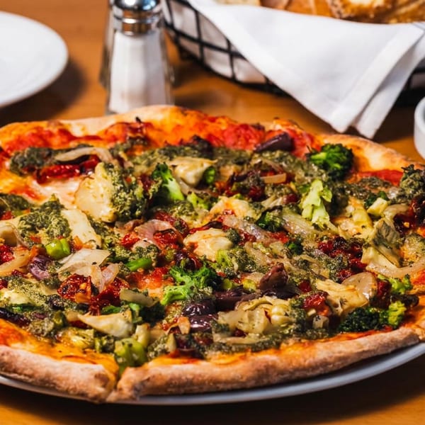 A whole vegetarian pizza with broccoli, artichokes, sun-dried tomatoes, and pesto on a wooden table.