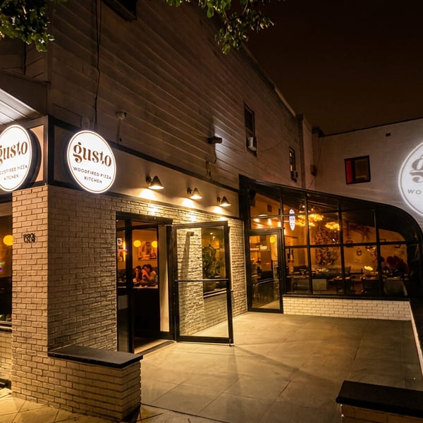 Exterior of Gusto Woodfired Pizza Kitchen at night, with illuminated signs and a projected logo.